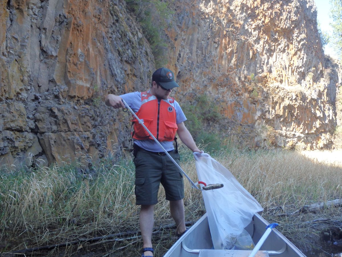 Lake Plastic Recovery: متطوع يرتدي سترة نجاة يجمع النفايات من شاطئ بحيرة ويضعها في زورق تحت منحدرات صخرية